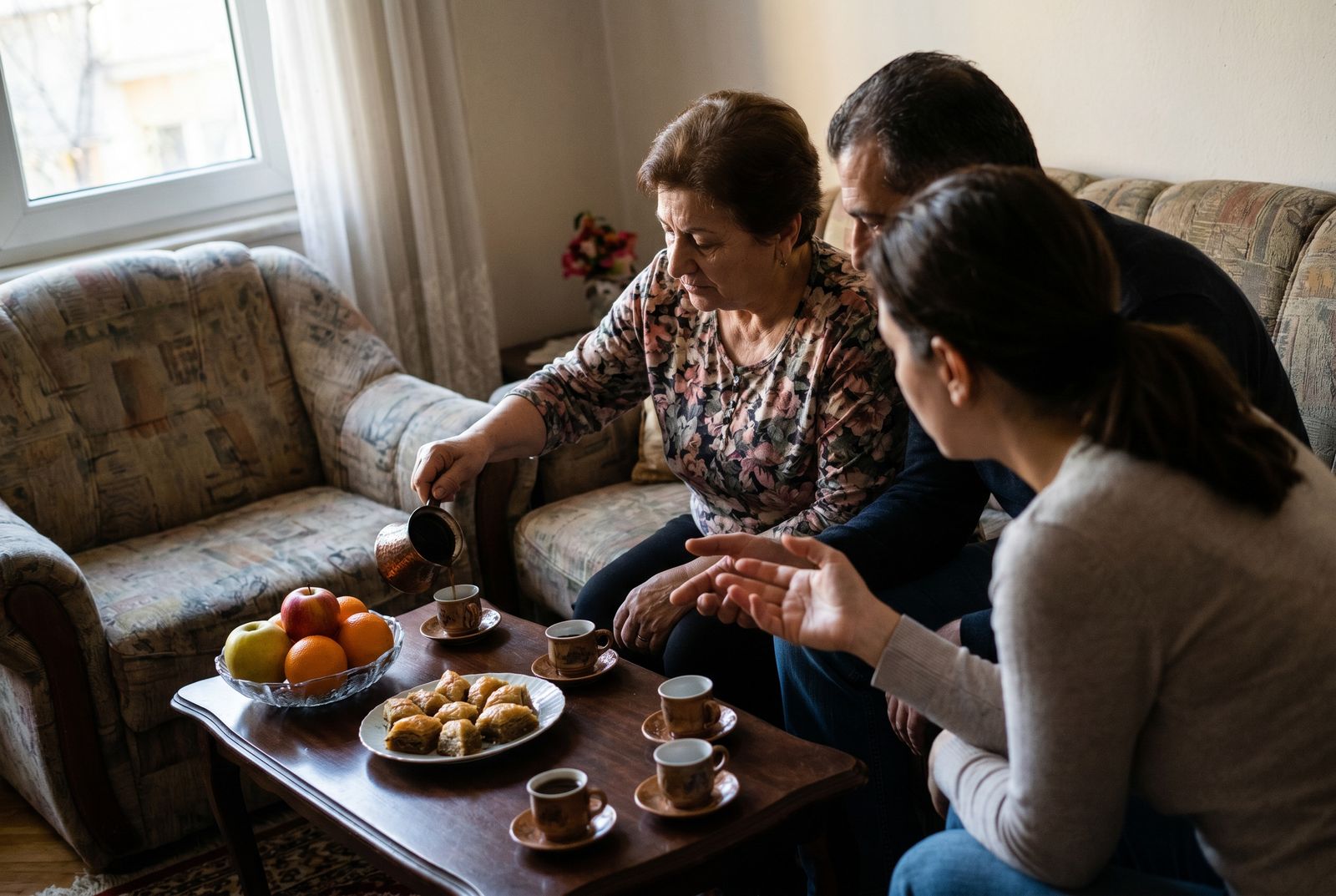 Tryezë e shtruar me kafe dhe ëmbëlsira në një dhomë ndenjeje shqiptare, disa persona ulur bashkë